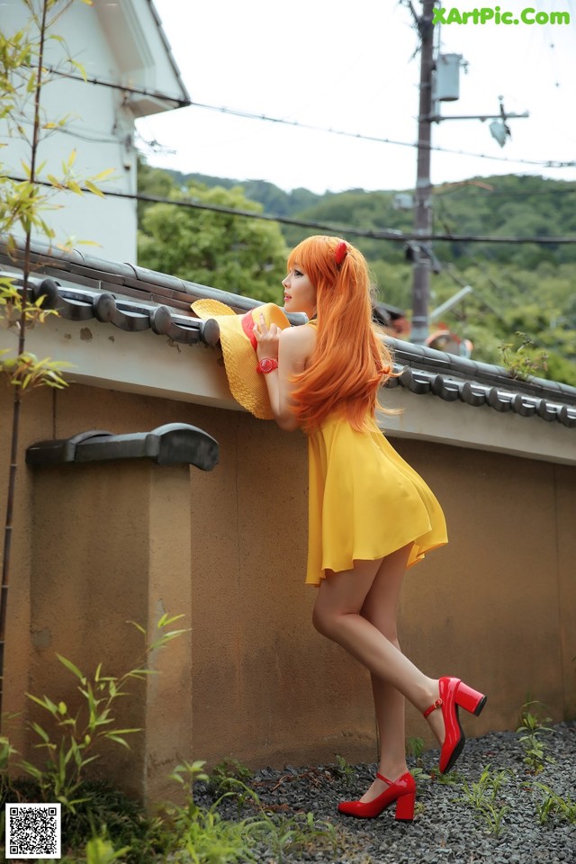 A woman in a yellow dress and red shoes leaning against a wall.