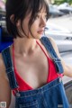 A woman in a red top and blue overalls leaning against a car.