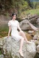 A woman in a white dress sitting on a rock by a river.